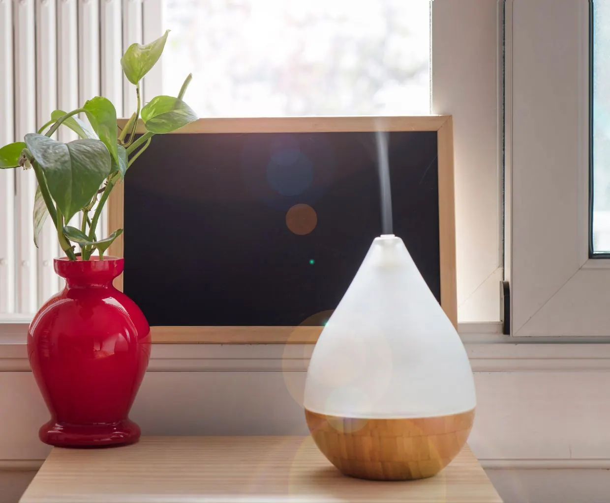 Teardrop-shaped white diffuser with a wooden base on a sunny windowsill next to a pothos plant in a red vase