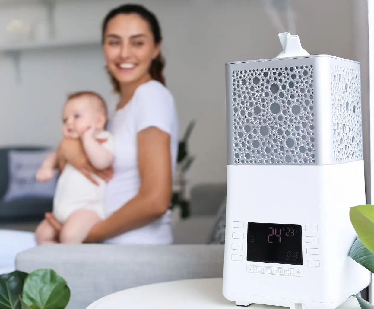 Smiling woman holding a baby in a living room, with a white humidifier releasing mist in the foreground