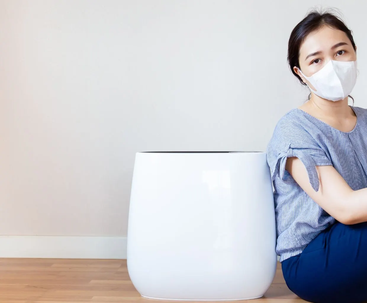 Woman wearing a white face mask sitting on a wooden floor next to a large white round air purifier
