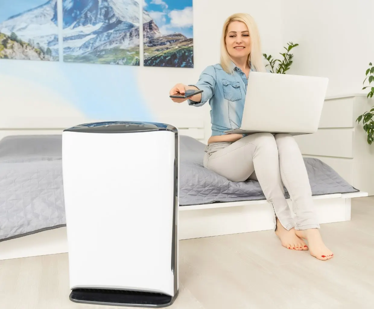 Woman with a laptop on a bed pointing a remote at a white floor-standing air purifier
