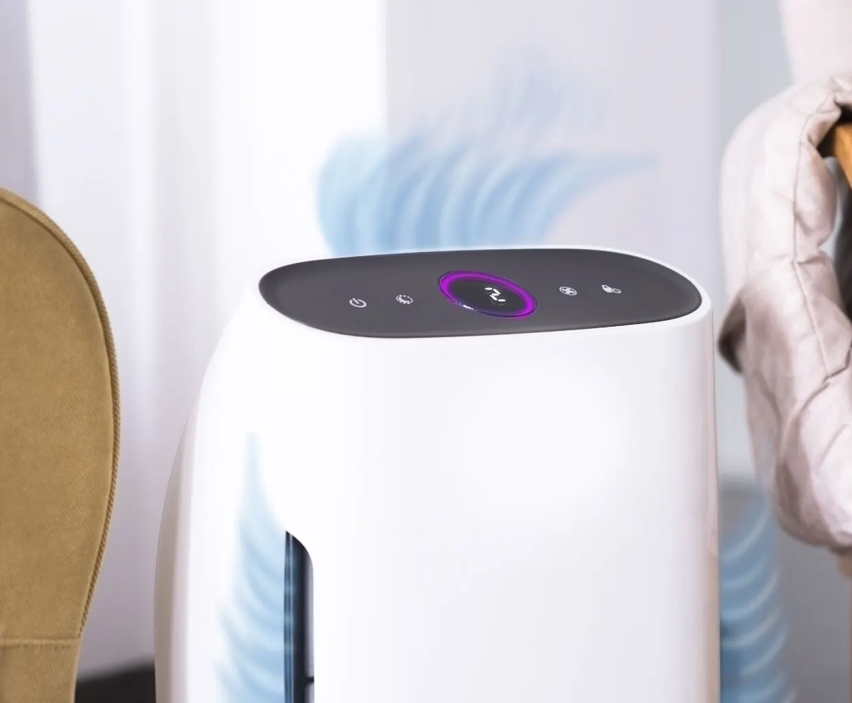Close-up of a white air purifier with a dark touch-control top and a purple display ring next to an armchair