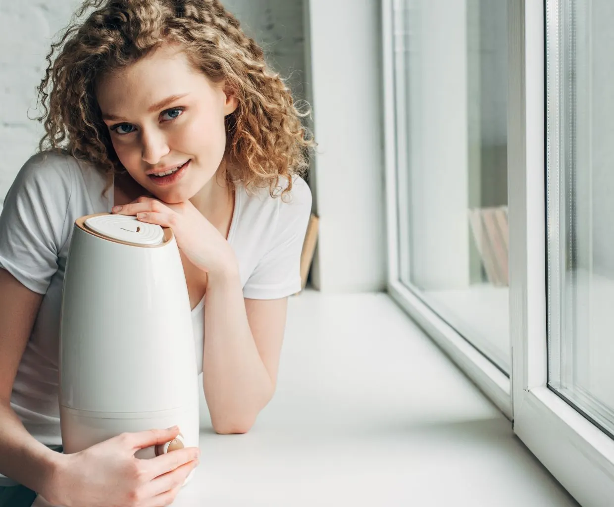 Woman with curly hair leaning on a small white tabletop appliance on a windowsill in a bright room