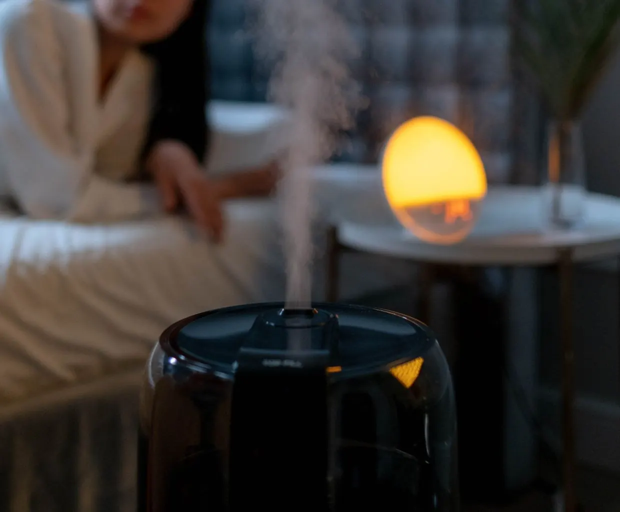 Humidifier releasing mist at night beside a bed, lit by a warm orange lamp