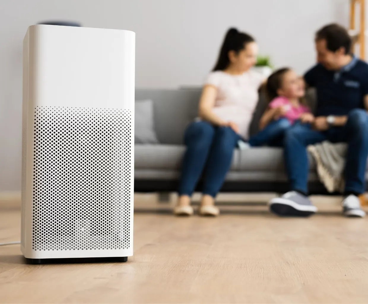 White air purifier with a perforated grille on a wood floor, with a family of three blurred on a gray sofa behind it