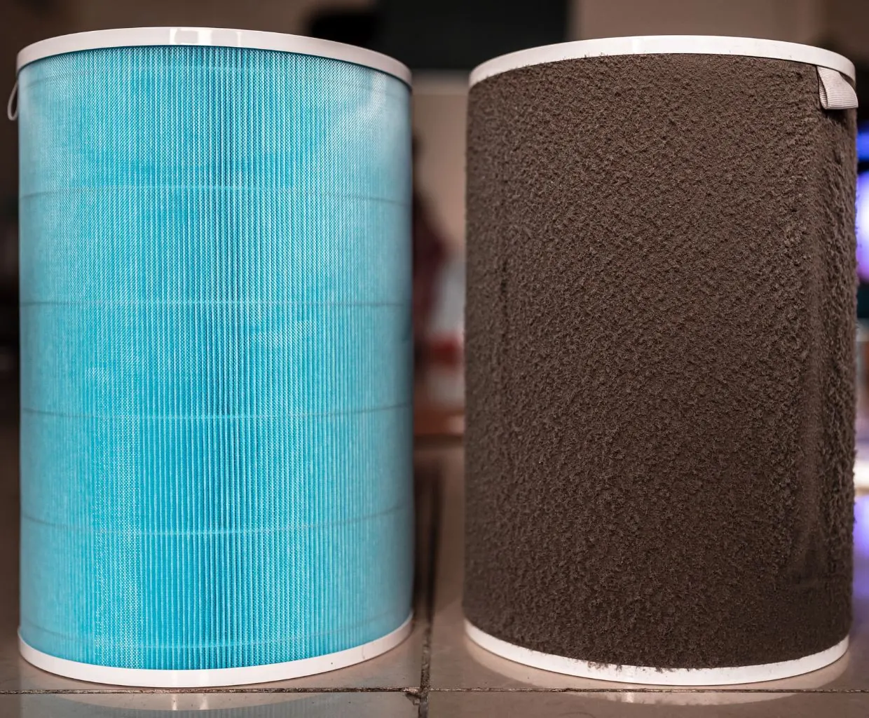 New blue pleated cylindrical air purifier filter next to a used filter coated in gray dust