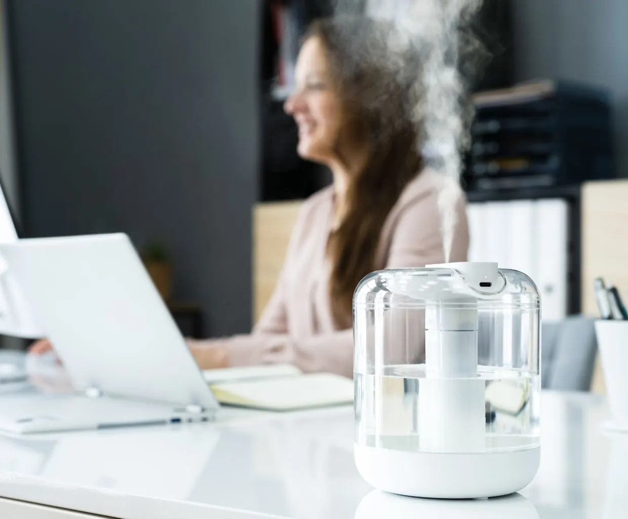 Clear-tank cool mist humidifier misting on a white office desk, with a woman working at a laptop behind it