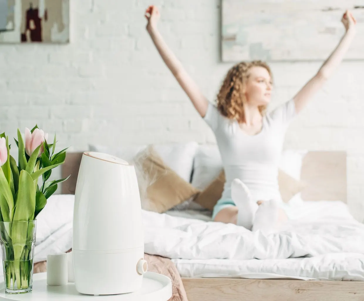 Woman stretching in bed behind a white humidifier and a vase of pink tulips on a bedside table