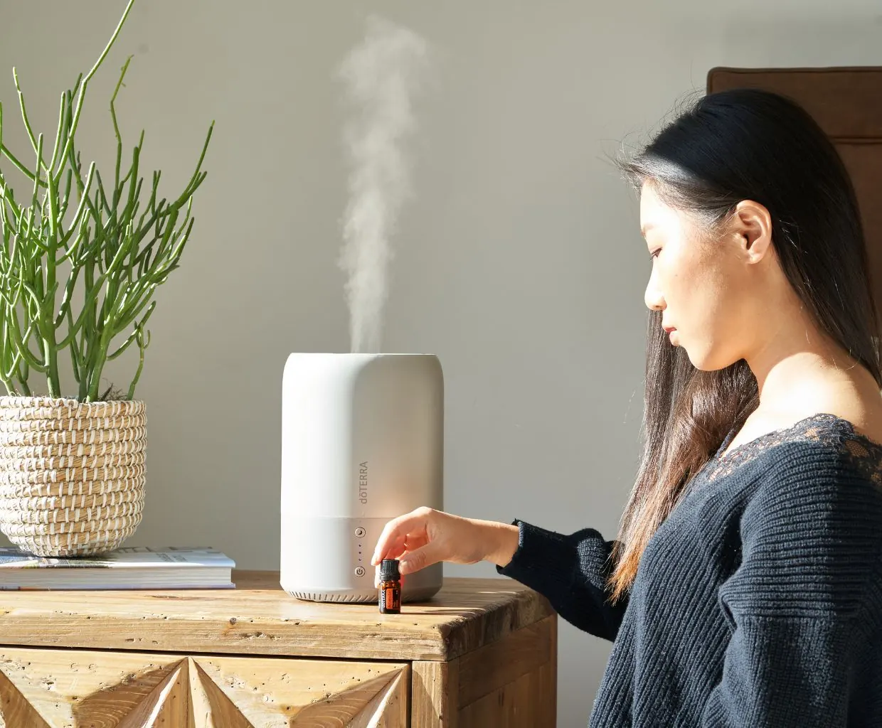 Woman setting a small bottle beside a white humidifier releasing mist on a wooden dresser next to a potted plant
