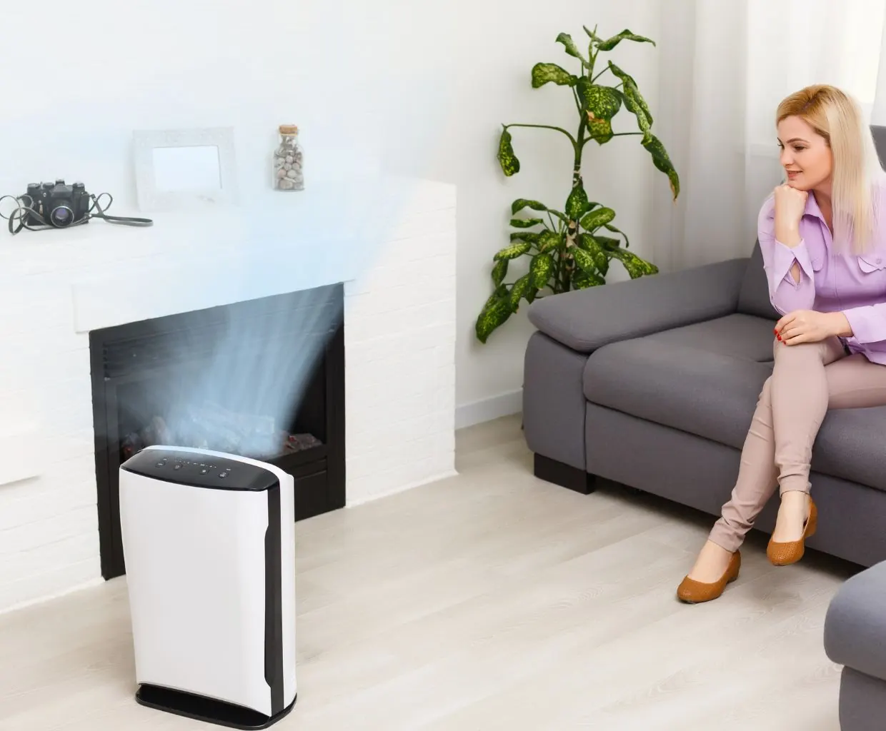 Woman on a gray sofa looking at a white tower air purifier standing in front of a fireplace