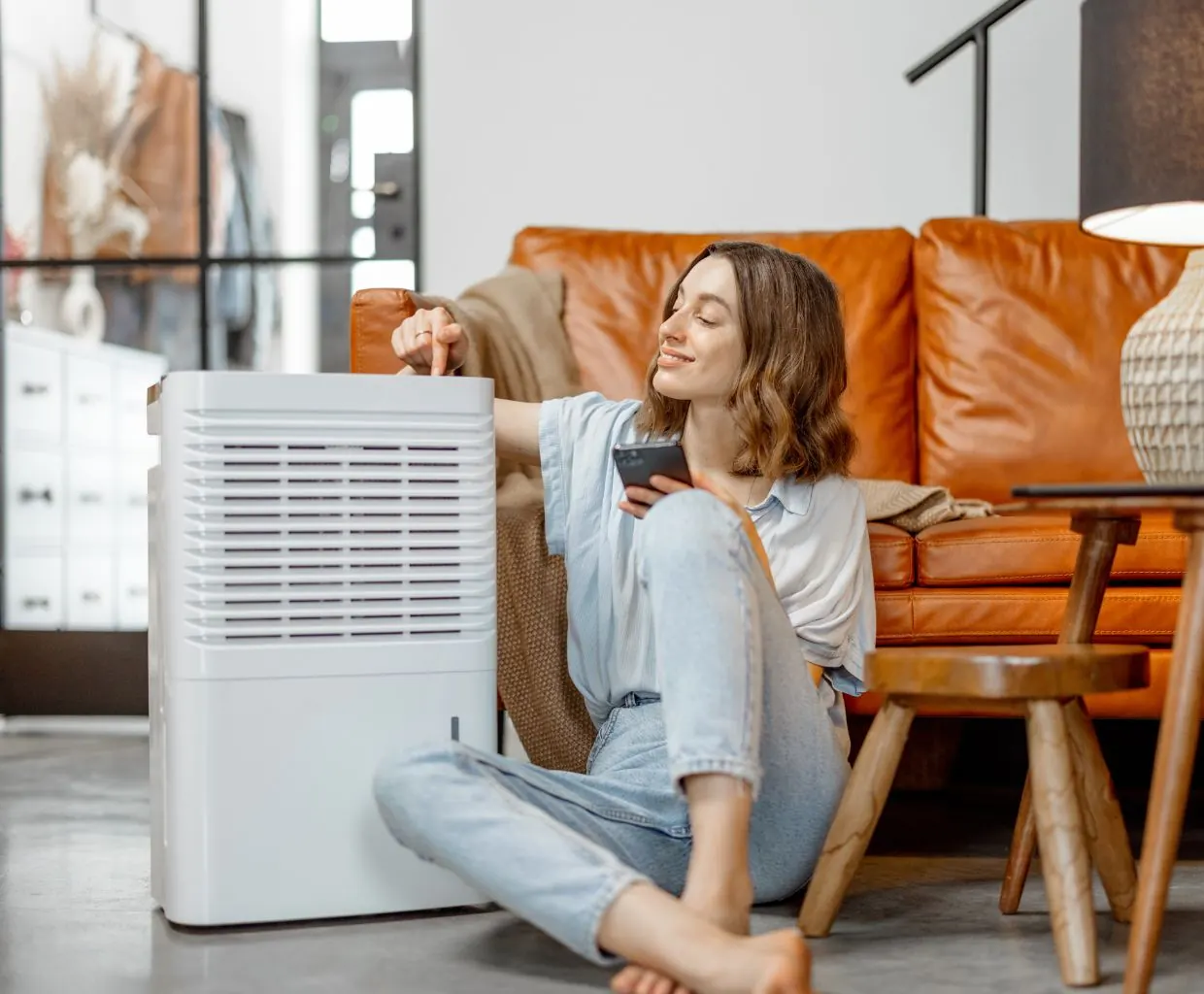 Woman sitting on the floor pressing a button on a large white air purifier next to an orange leather sofa