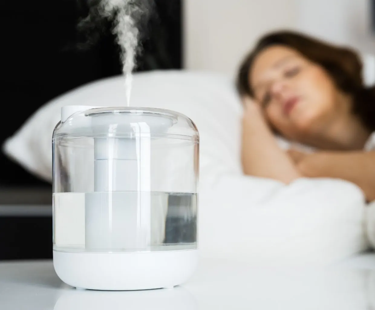 Clear-tank humidifier releasing mist on a nightstand while a woman sleeps in bed behind it
